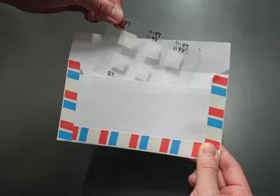 One hand holds a white envelope with a blue and red striped border, while a second hand places a clear sheet of plastic inside with small squares of paper on it